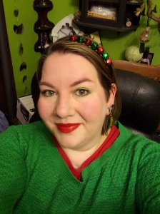 Picture of me wearing a green sweater with a red undershirt, a red and green jingle bell headband, christmas tree earrings, green eyeshadow and red lipstick. 